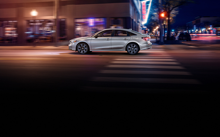 2026 Honda Civic Sedan Sport Touring Hybrid in Solar Silver Metallic profile driving on a city street (Compact Sedan).