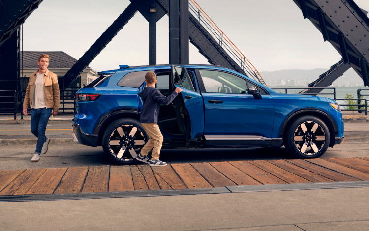 2026 Honda Prologue Elite in Pacific Blue Metallic, parked along the curb under a black industrial drawbridge for a fun family day.