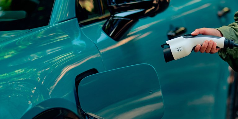 2026 Honda Prologue Elite in North Shore Pearl, parked in redwoods, with a woman preparing to plug the portable charger into the vehicle.